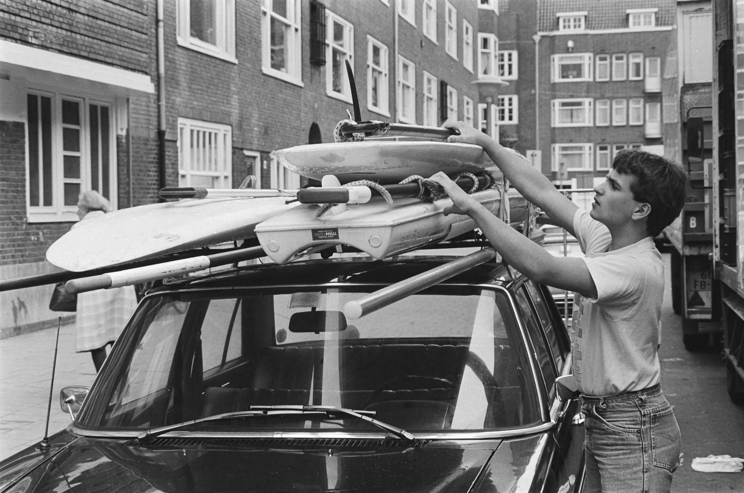 People lifting a sailboard onto the roof of a car in a street