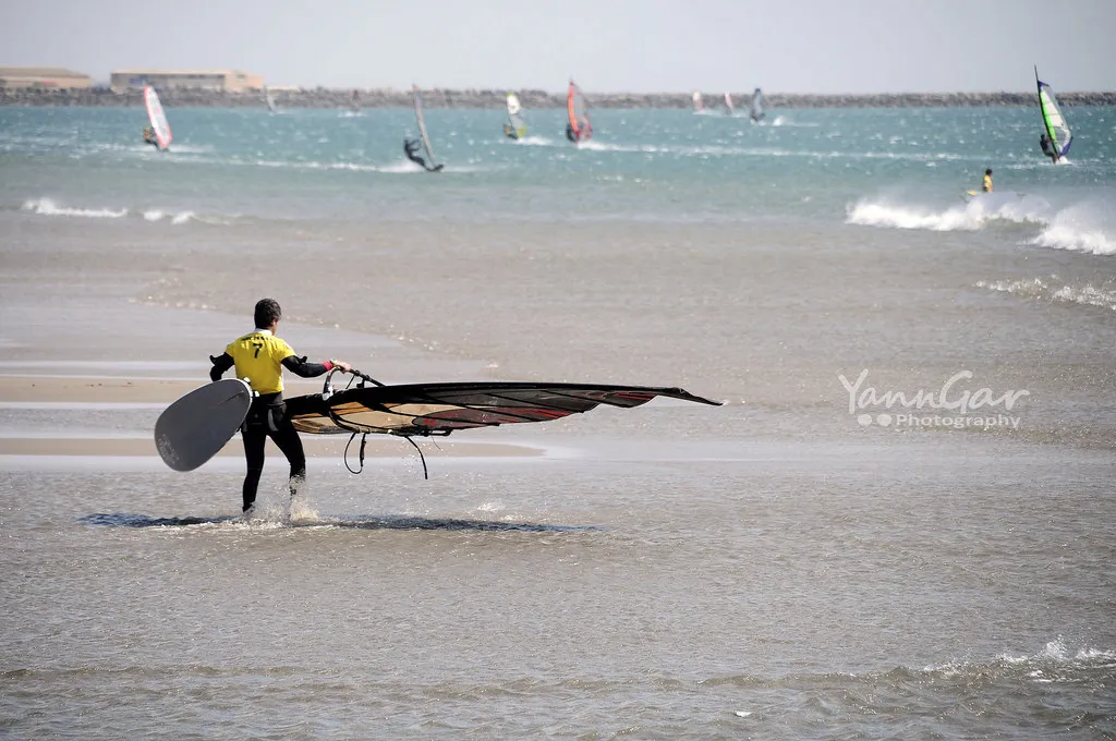A windsurfer carrying his board and sail into the sea, with other windsurfers behind
