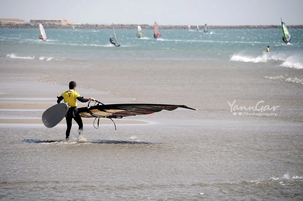 A windsurfer carrying his board and sail into the sea, with other windsurfers behind