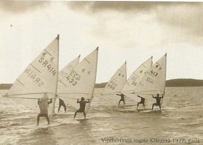 Six windsurfers with numbered sails standing on their boards in shallow water