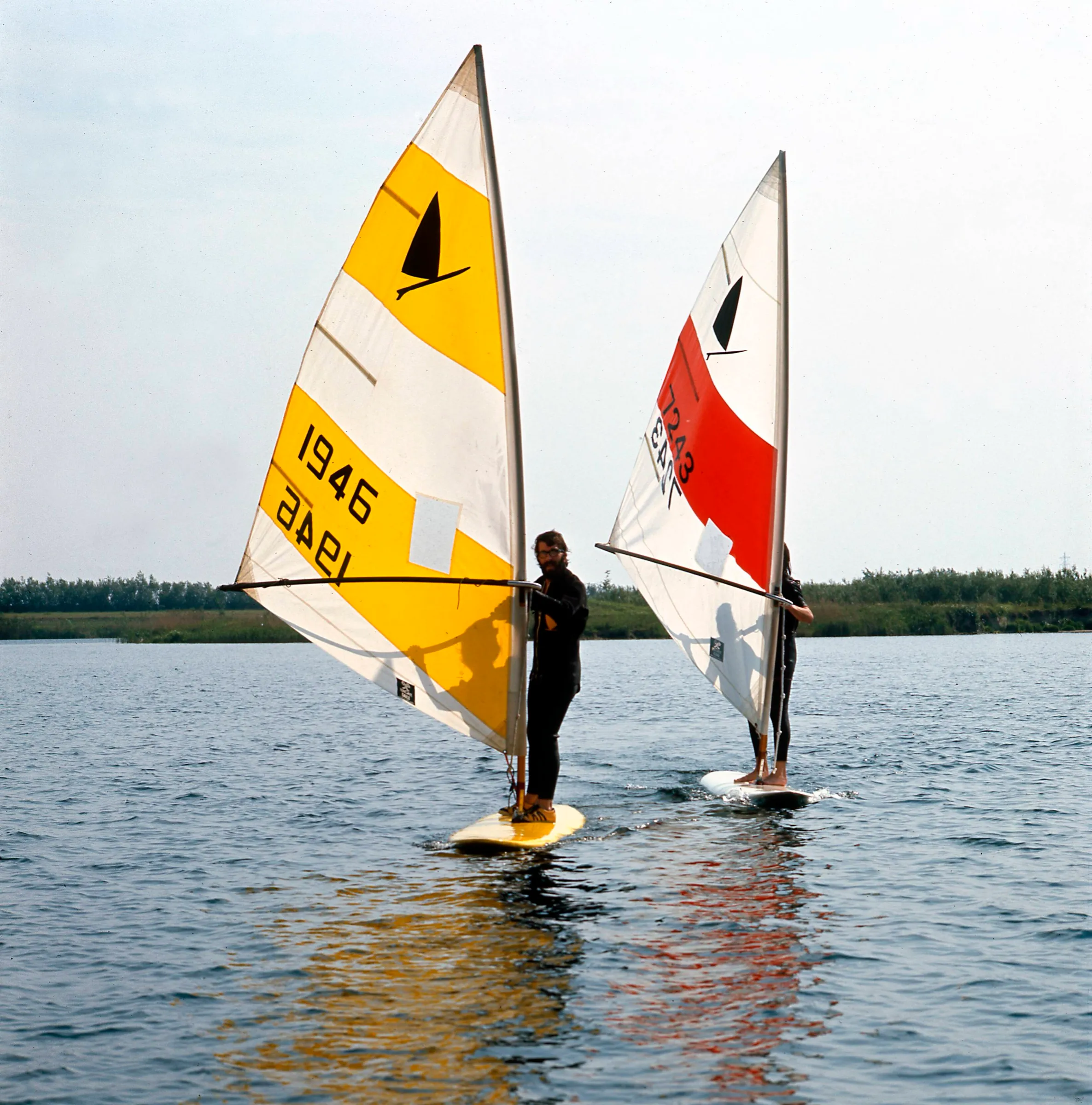 Two windsurfers with yellow and red sails on calm water